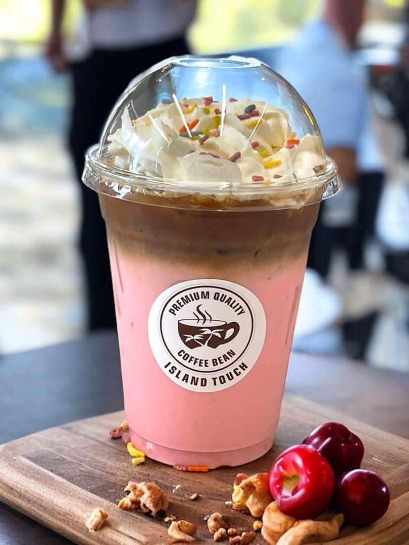 Pink and brown layered iced coffee with whipped cream and sprinkles, next to cherries and nuts.